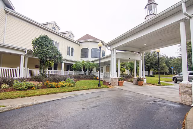 Exterior view of a senior living facility with a covered entrance driveway, landscaped garden with shrubs and flowers, and a two-story building with large windows and white railings.