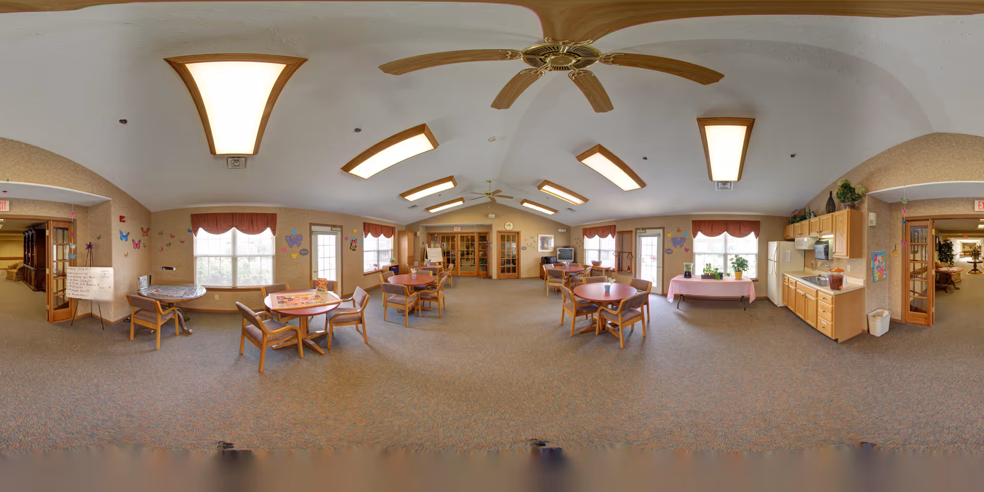 A spacious, well-lit common area in a senior living facility with multiple round tables and chairs arranged for group activities or dining. The room features large windows with valances, ceiling fans, overhead lights, and a kitchenette area with cabinets, a refrigerator, and a microwave. Walls are decorated with colorful butterfly and heart decals, and there is a whiteboard with writing near the entrance. The room has carpeted flooring and doors leading to adjacent hallways.