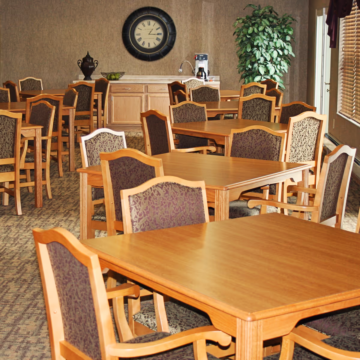 A dining room with multiple wooden tables and chairs featuring patterned upholstery. A large round clock hangs on the wall above a wooden cabinet, and a tall green plant is placed in the corner near a window with blinds.