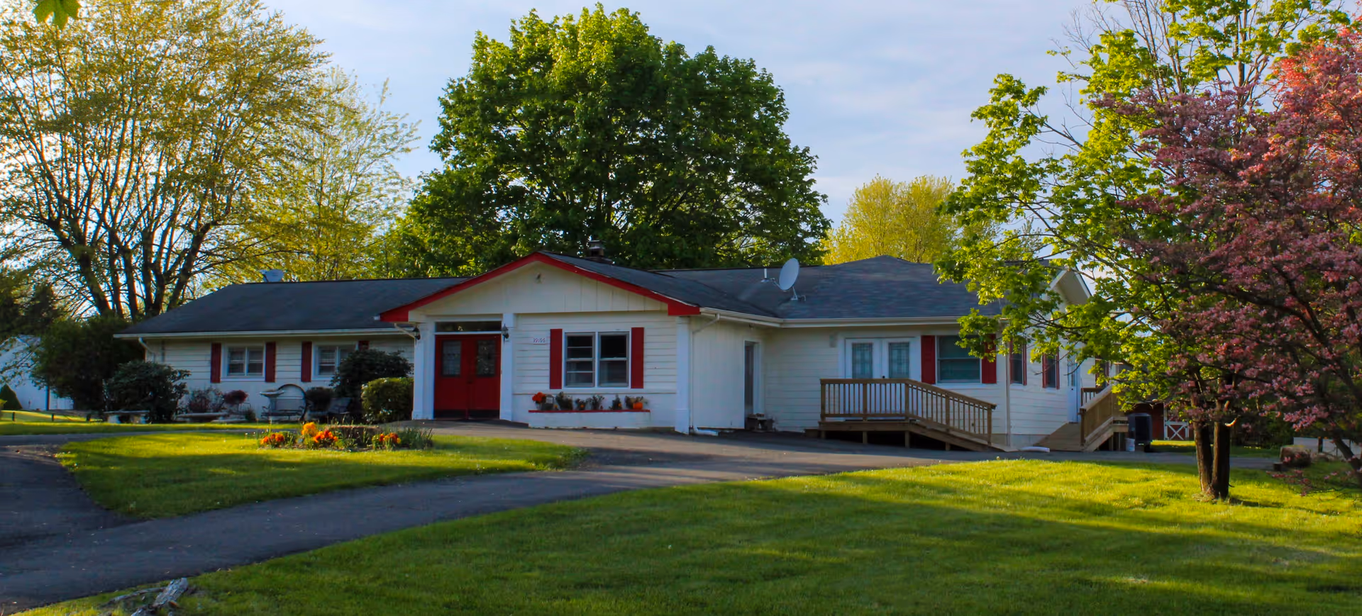 Single-story white assisted living building with red trim, a front lawn, driveway, and trees.