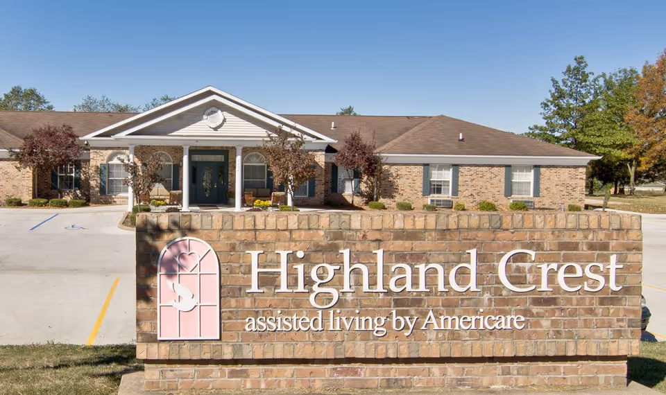 Exterior view of Highland Crest assisted living facility showing a brick building with a peaked roof, white columns at the entrance, and a large brick sign in the foreground with the facility name and logo.
