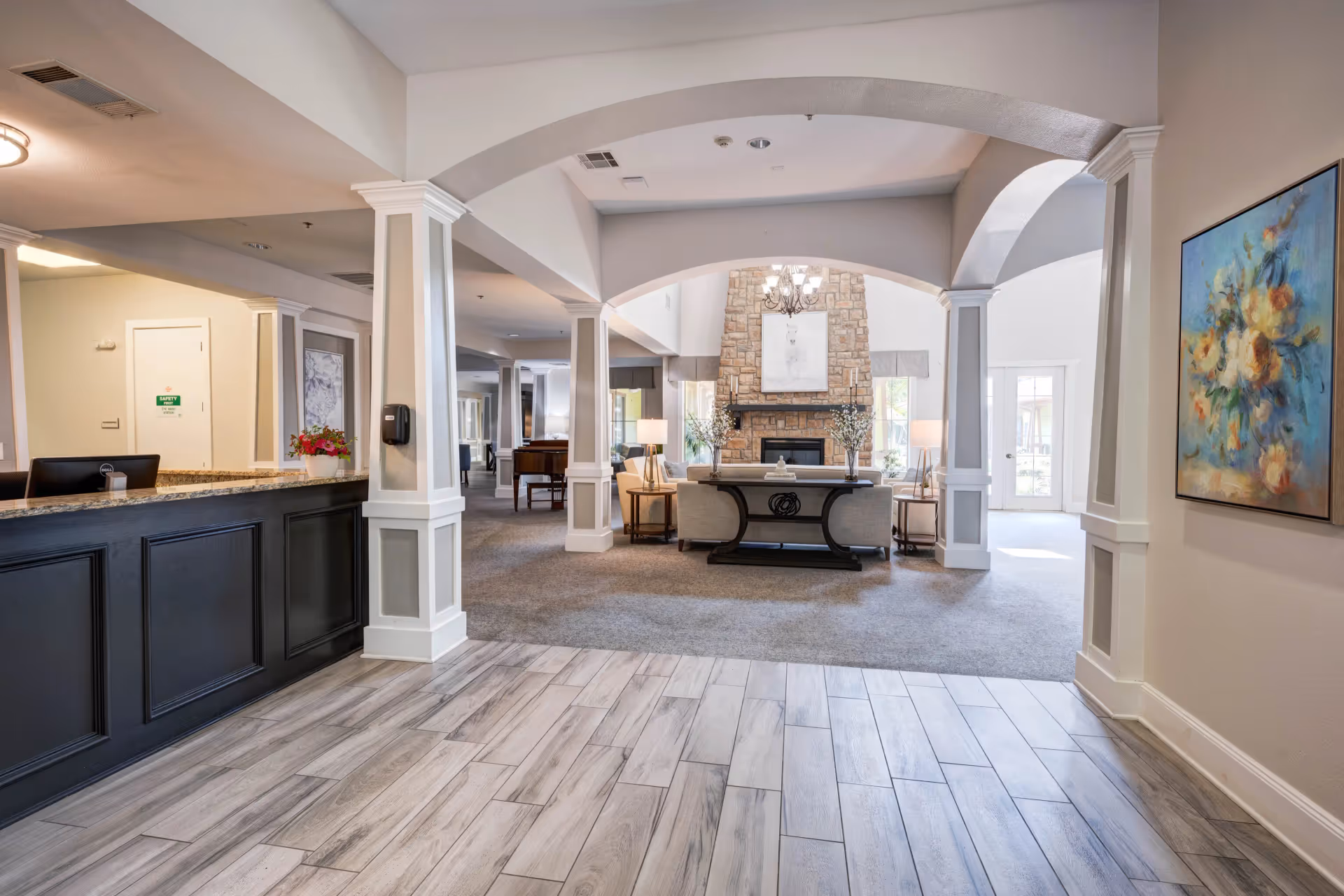 Interior view of a senior living facility lobby with a reception desk on the left, a seating area with sofas and lamps in front of a stone fireplace, and a painting of flowers on the right wall. The floor near the entrance is tiled, transitioning to carpet in the seating area. The space is well-lit with natural light coming through glass doors in the background.
