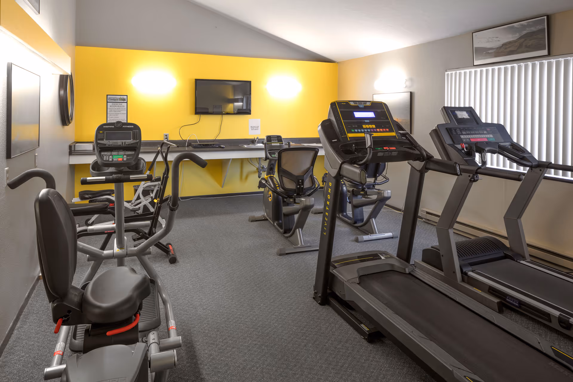 A small fitness room with exercise equipment including two treadmills, two recumbent stationary bikes, and one upright stationary bike. The room has a yellow accent wall with a mounted TV and a long desk with a chair underneath. There are framed pictures on the walls and a window with vertical blinds.