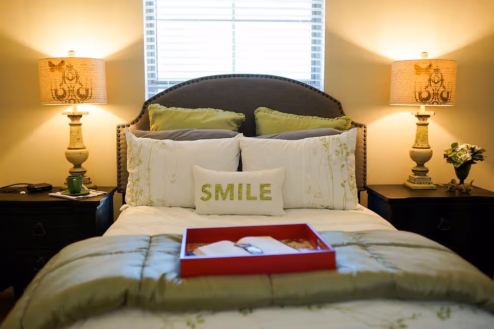 Neatly made bed in a softly lit bedroom with decorative pillows (one reading "SMILE"), matching nightstands and lamps.