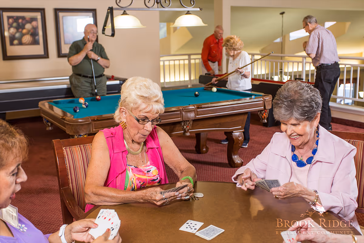 A group of elderly people enjoying recreational activities in a common area. Three women are seated at a table playing cards, while three men and one woman are playing pool in the background. The room has warm lighting and framed artwork on the walls.