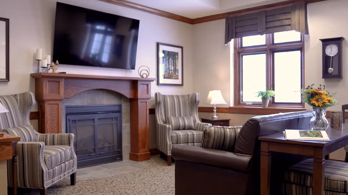 Comfortable common living room with a fireplace and wall-mounted TV, striped armchairs, a leather sofa, and a table with flowers by a window.