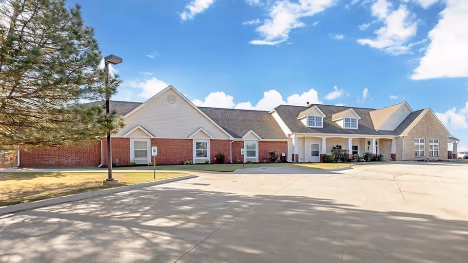 Exterior view of a single-story senior living facility building with a combination of brick and light-colored siding, multiple windows, a covered entrance, and a large paved parking area in front under a partly cloudy blue sky.