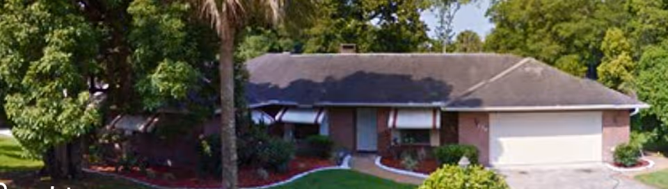 Single-story brick house with a dark roof, white garage door, and several windows with awnings. The house is surrounded by green trees and bushes, with a well-maintained lawn and a concrete driveway.