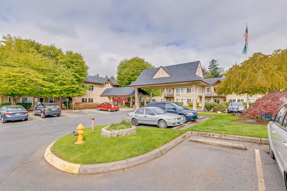 Exterior view of Solstice Senior Living At Normandy Park showing a parking lot with several cars, a covered entrance, green trees, and a flagpole with an American flag.