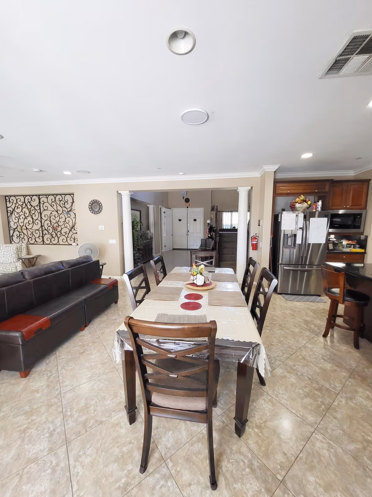 Interior view of a residential assisted living home showing a dining area with a rectangular table covered with a tablecloth and six chairs. To the left, there is a dark brown leather sofa and decorative wall art. In the background, there is a hallway leading to a white front door. To the right, a kitchen area is visible with a stainless steel refrigerator, wooden cabinets, and a bar stool.
