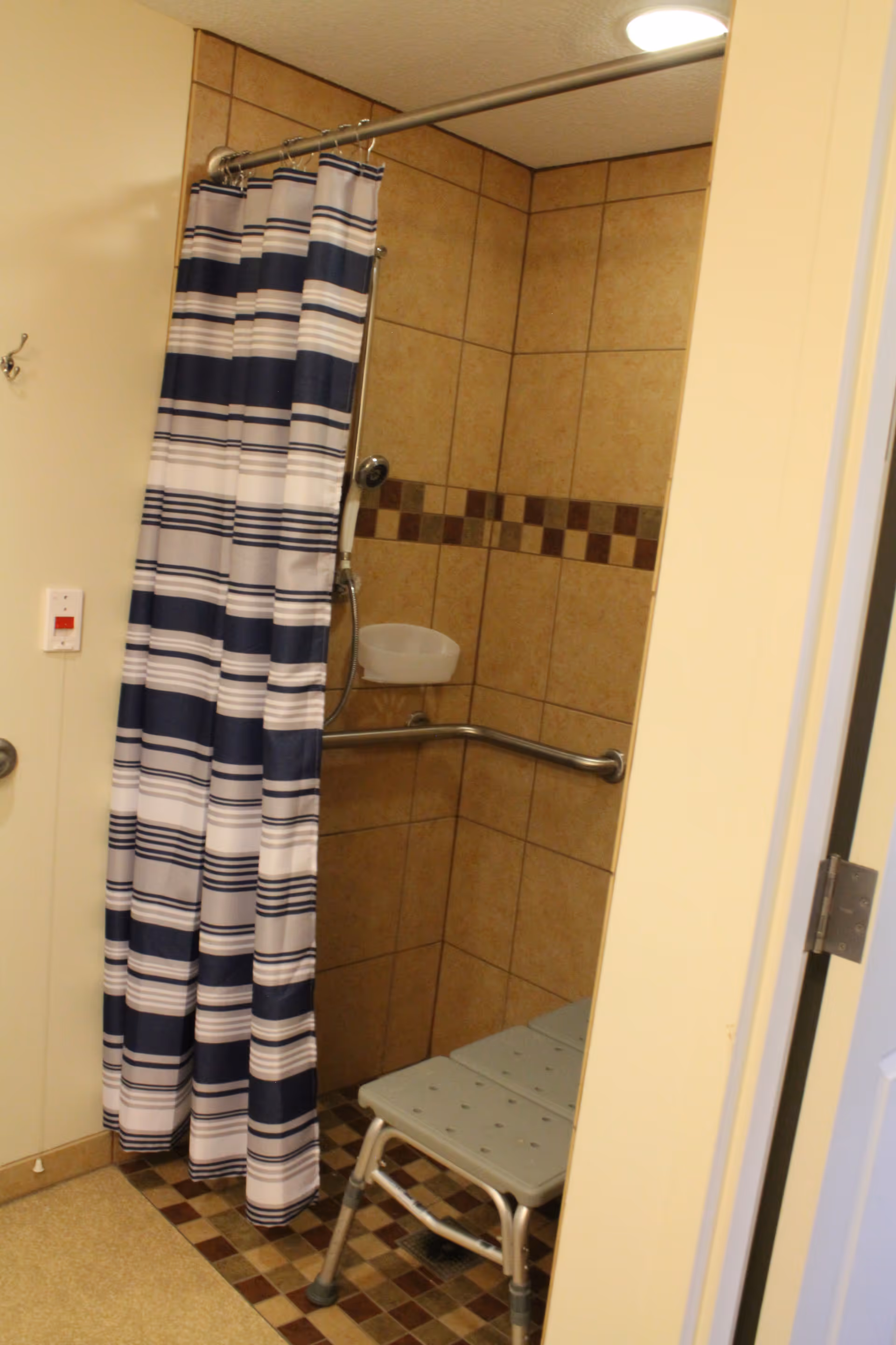 Tiled shower stall with a striped shower curtain, grab bar and a shower chair.