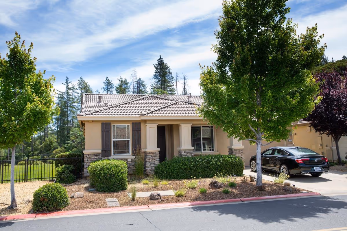 Single-story residential building with beige walls, stone accents, and a tiled roof. The front yard has neatly trimmed bushes, small trees, and a driveway with a black car parked. The sky is partly cloudy with blue patches visible.