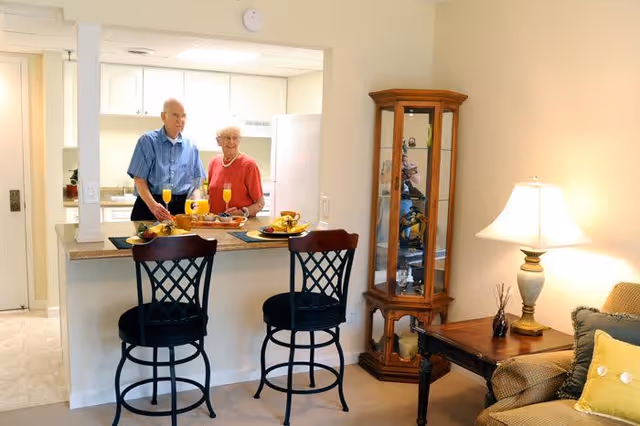 An elderly couple standing behind a kitchen counter with two glasses of orange juice and plates of food. The kitchen counter has two high chairs in front of it. To the right, there is a wooden display cabinet with glass doors and a side table with a lamp and some decorative items. A beige couch with cushions is partially visible on the right side.