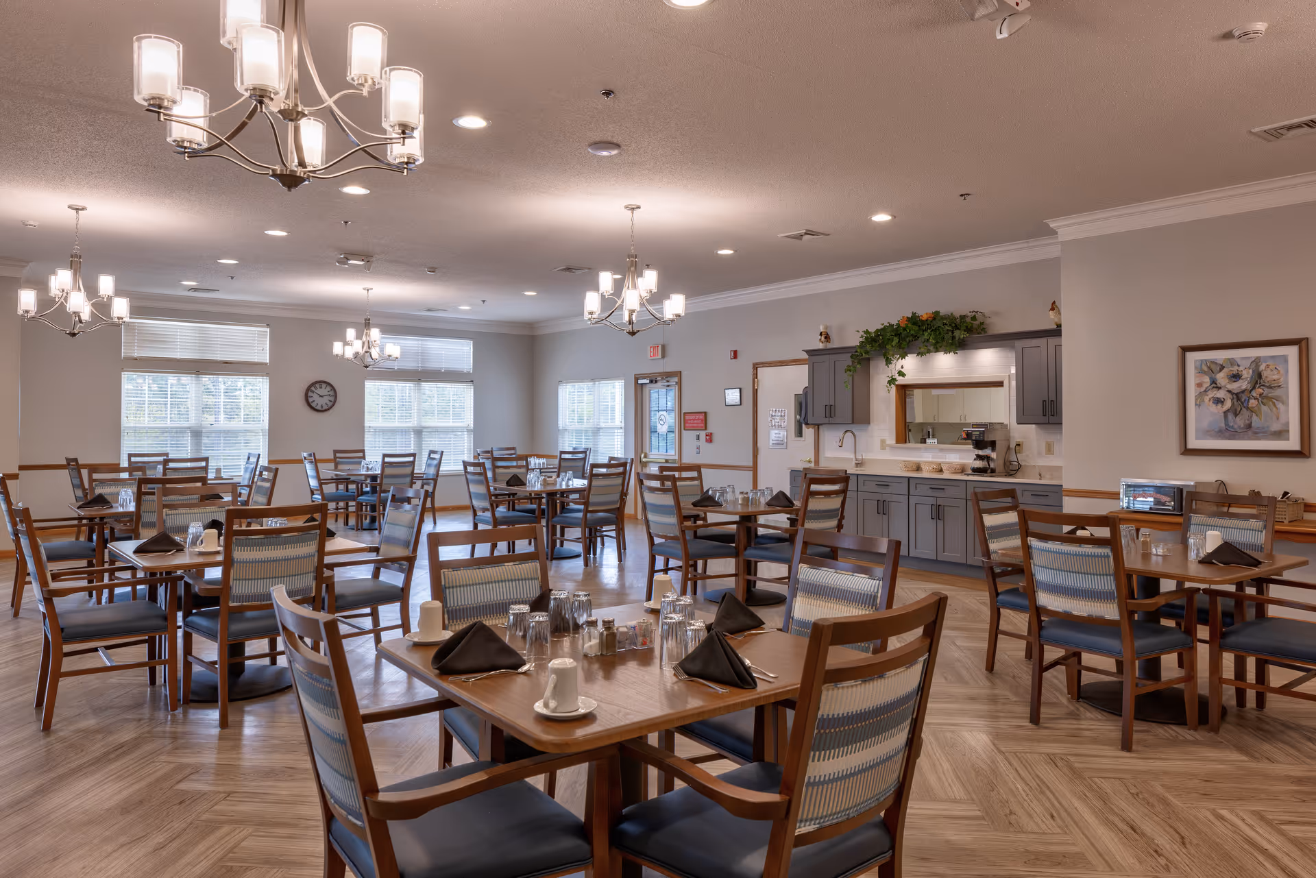 A spacious dining room with multiple wooden tables and chairs arranged neatly. Each table is set with black napkins, glasses, cups, and silverware. The room features large windows with blinds, modern chandeliers hanging from the ceiling, and a counter area with cabinets and a coffee machine. The walls are painted light gray, and there is a framed floral painting on one wall.