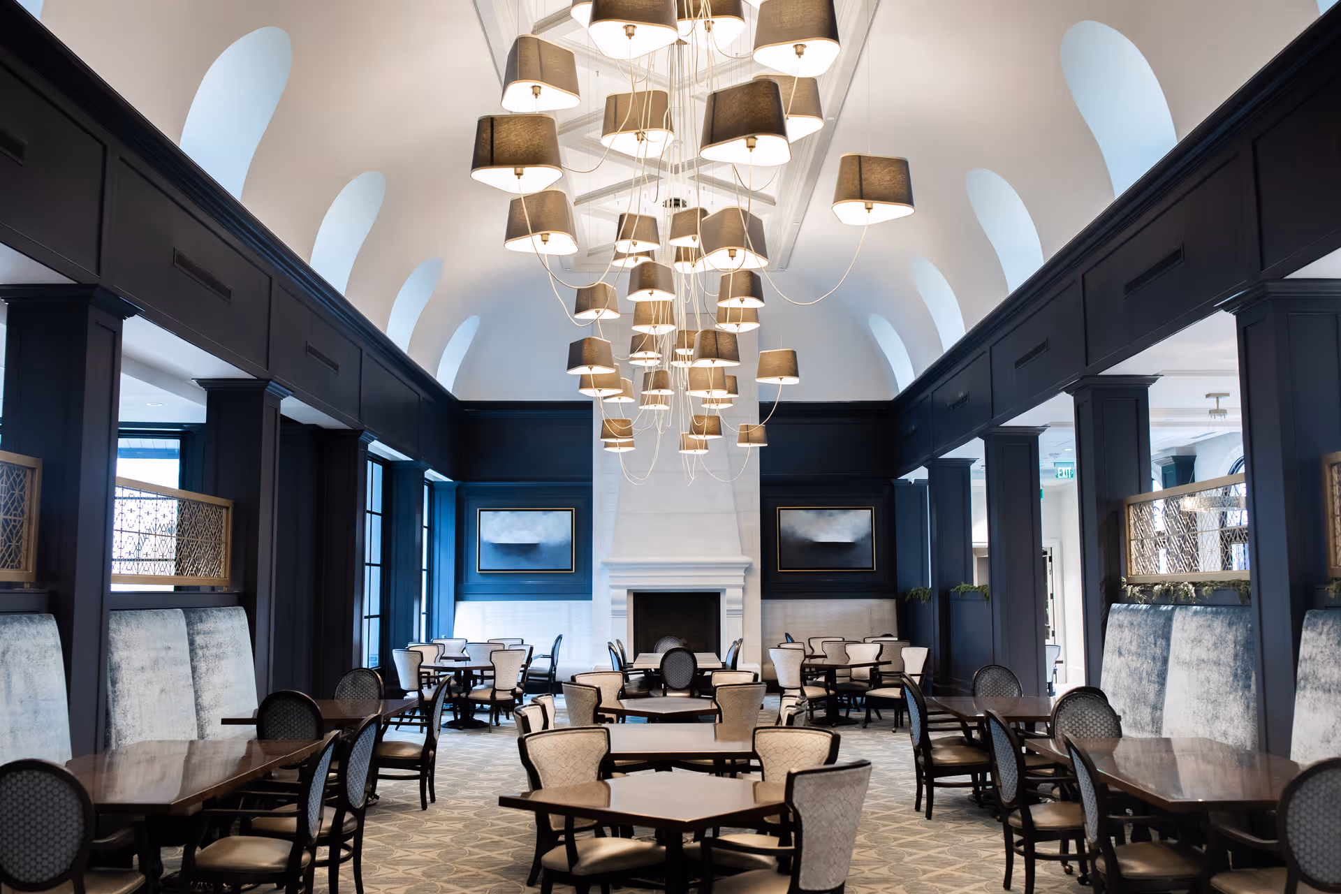 Elegant dining room with a high vaulted ceiling and clustered pendant lights above rows of tables and chairs facing a central fireplace.