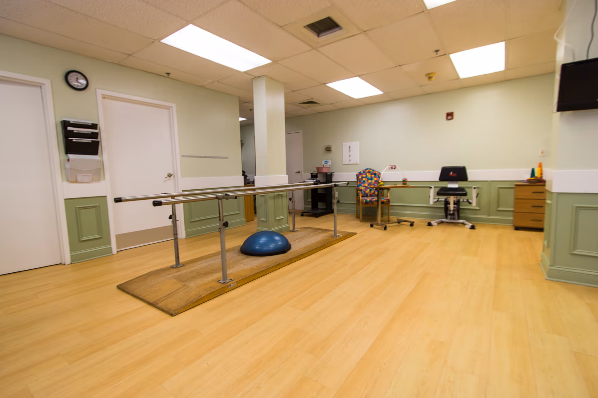 A rehabilitation room with parallel bars for physical therapy, a blue balance dome on a wooden platform, a colorful patterned chair, a black office chair, a small table with therapy equipment, light green walls with white trim, and light wood flooring.