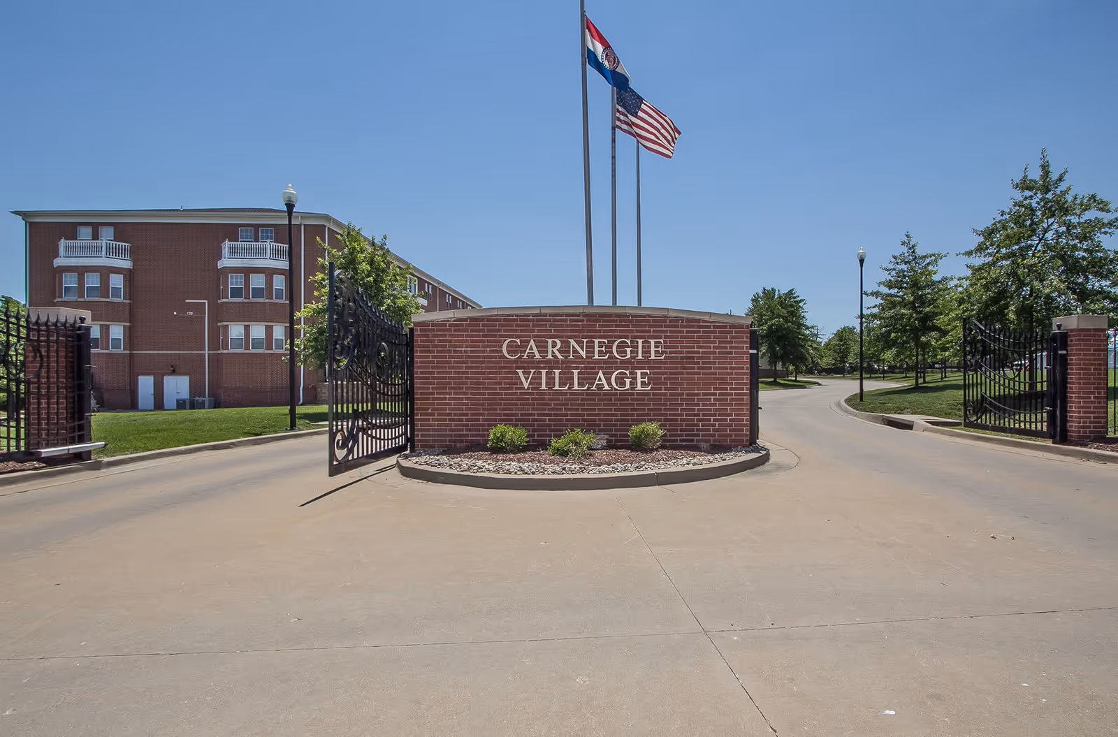 Entrance to Carnegie Village Senior Living Community featuring a brick sign with the community name, two flagpoles with American and state flags, black wrought iron gates, a paved driveway, and a multi-story brick building in the background under a clear blue sky.