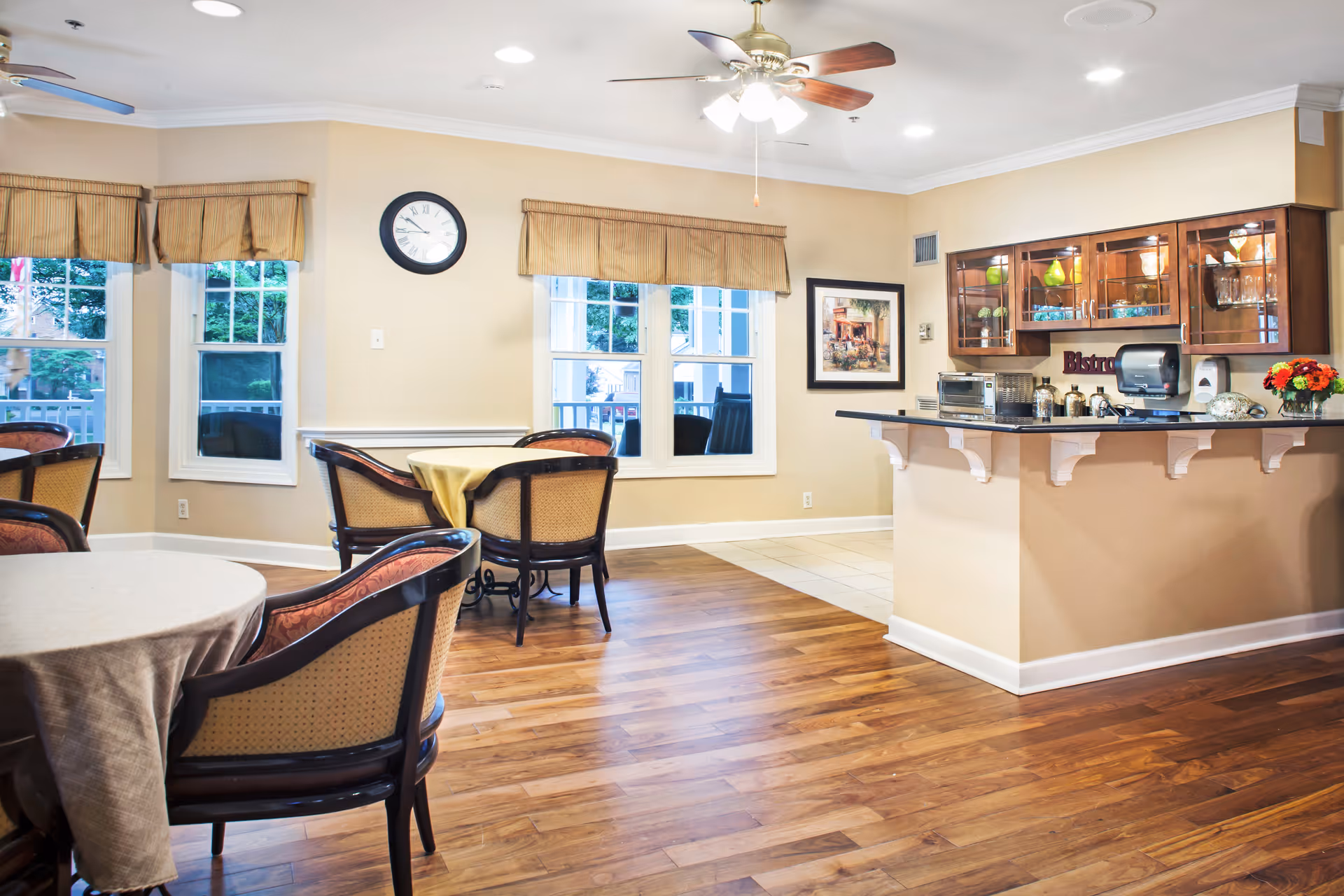 A bright and clean dining area in a senior living facility with wooden floors, round tables covered with tablecloths, and cushioned chairs. The room features large windows with beige valances, a ceiling fan with lights, a wall clock, and a small kitchen counter area with glass cabinets displaying decorative items and kitchen appliances.