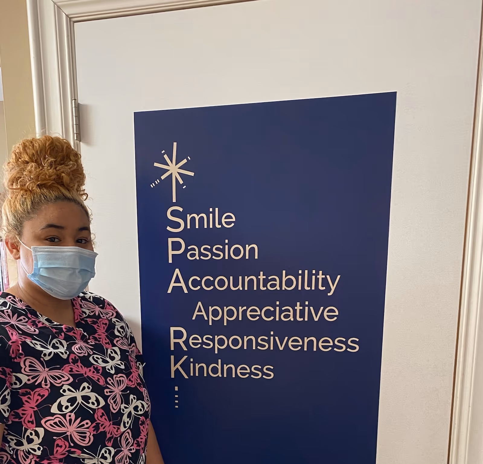 A woman wearing a face mask and a butterfly-patterned scrub top stands next to a blue poster on a white door. The poster displays the acronym SPARK with the words Smile, Passion, Accountability, Appreciative, Responsiveness, and Kindness listed vertically.