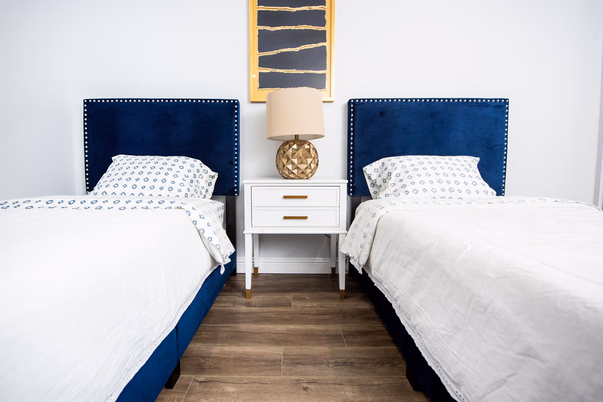 A bedroom with two twin beds featuring dark blue upholstered headboards with silver nailhead trim. Each bed has white bedding with a subtle pattern on the pillows and sheets. Between the beds is a white nightstand with two drawers and gold handles, topped with a decorative gold lamp with a beige lampshade. Above the nightstand hangs a framed abstract art piece with a gold frame. The floor is wooden with a medium brown finish.