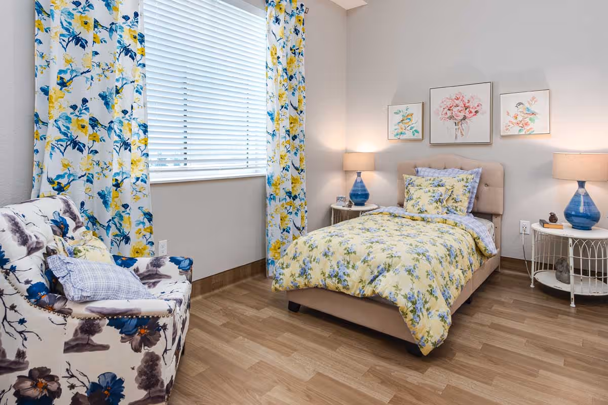 A cozy bedroom with a single bed dressed in yellow floral bedding. There are two blue lamps on white side tables on either side of the bed. Above the bed, three framed pictures of birds and flowers hang on the wall. A floral-patterned armchair is positioned near a window with white blinds and yellow and blue floral curtains. The room has wooden flooring and light-colored walls.