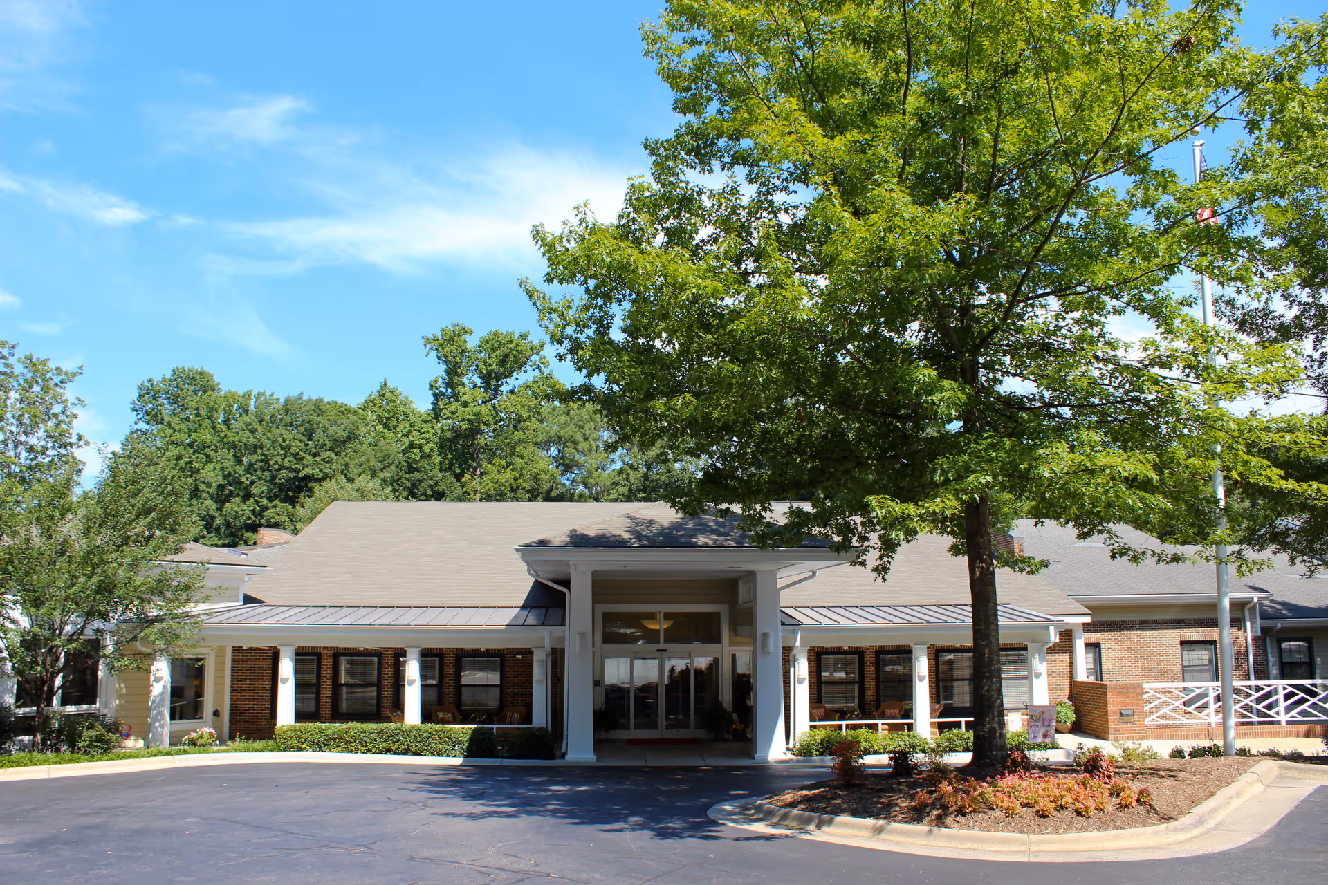 Front exterior view of a single-story senior living facility building with a covered entrance, surrounded by trees and landscaping under a clear blue sky.