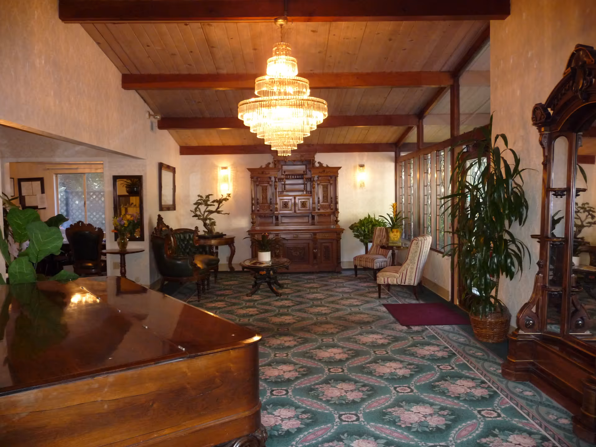 A decorative assisted-living lobby with a large chandelier, antique wooden furniture, potted plants, and patterned carpet.