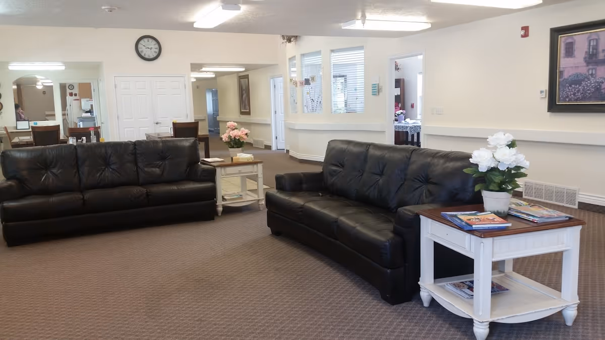 A senior living facility common area with two black leather sofas facing each other, a white side table with a potted white flower and magazines, another side table with a pink flower, and a hallway leading to other rooms. The walls are white, and there is a clock above a set of double doors in the background.