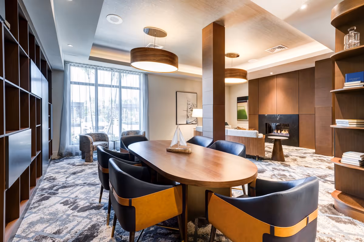 A modern and cozy living room and dining area with a wooden oval dining table surrounded by black and brown chairs. The room features large windows with sheer curtains, a fireplace with a dark surround, built-in wooden bookshelves, and contemporary ceiling lights. The carpet has a patterned design, and there are additional seating areas with armchairs near the windows.