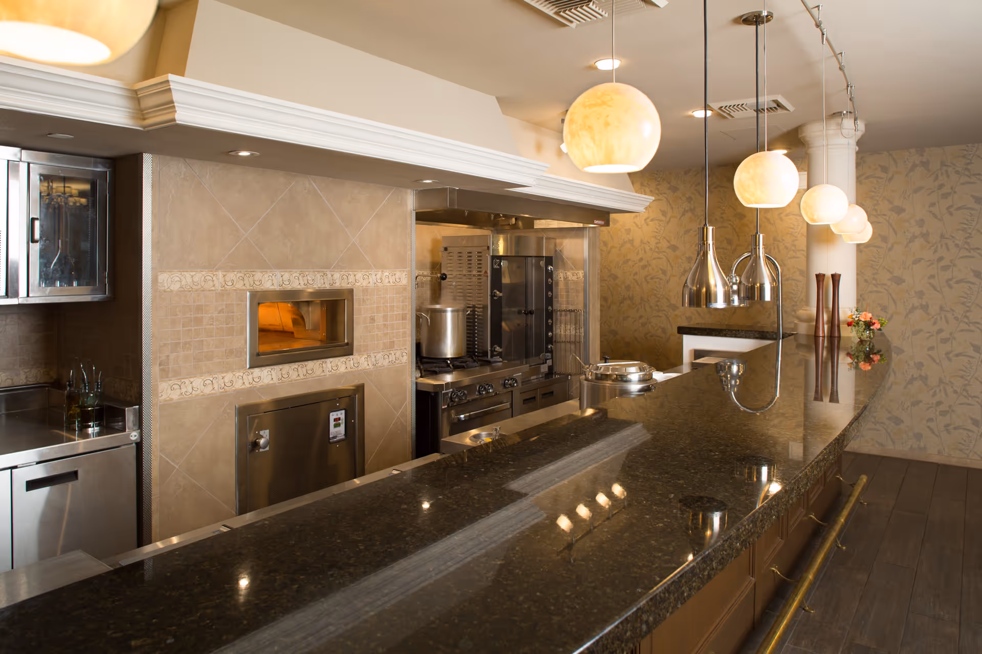 A modern kitchen area with a long dark granite countertop, hanging pendant lights, stainless steel appliances including an oven and stove, and a wood-fired pizza oven built into a tiled wall. The background features floral wallpaper and a small flower arrangement on the counter.