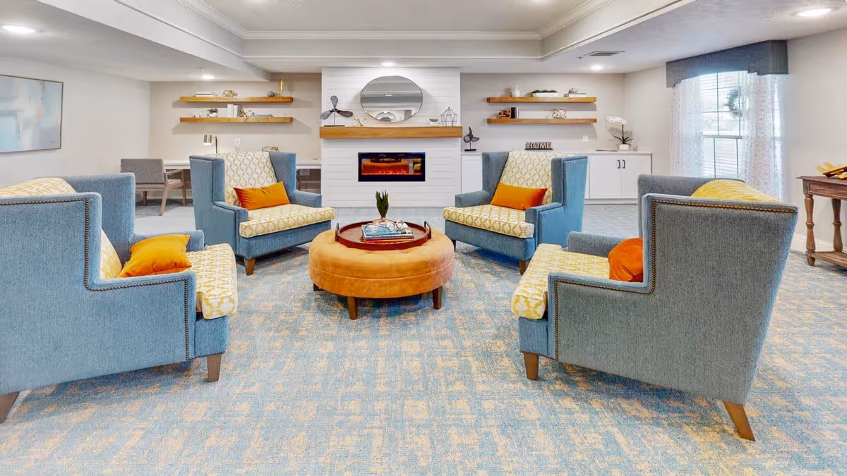 A cozy living room with four blue armchairs featuring yellow patterned cushions and orange throw pillows arranged around a round, tan ottoman. The room has a light blue patterned carpet, a white fireplace with a wooden mantel, a round mirror above the fireplace, and floating wooden shelves with decorative items. There is a window with sheer curtains and a valance on the right side, and a small wooden table with a lamp in the background.