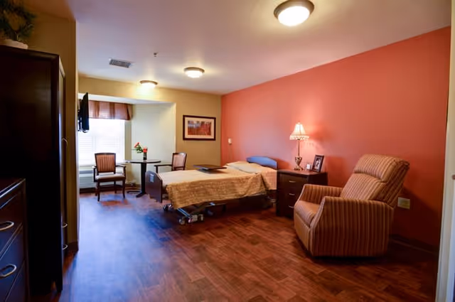 A cozy senior living bedroom with a single hospital-style bed covered with a beige blanket, a wooden nightstand with a lamp and framed photos, a striped armchair, two chairs and a small round table near a window with blinds and a valance, and warm wood flooring. The walls are painted in a combination of beige and muted red tones.