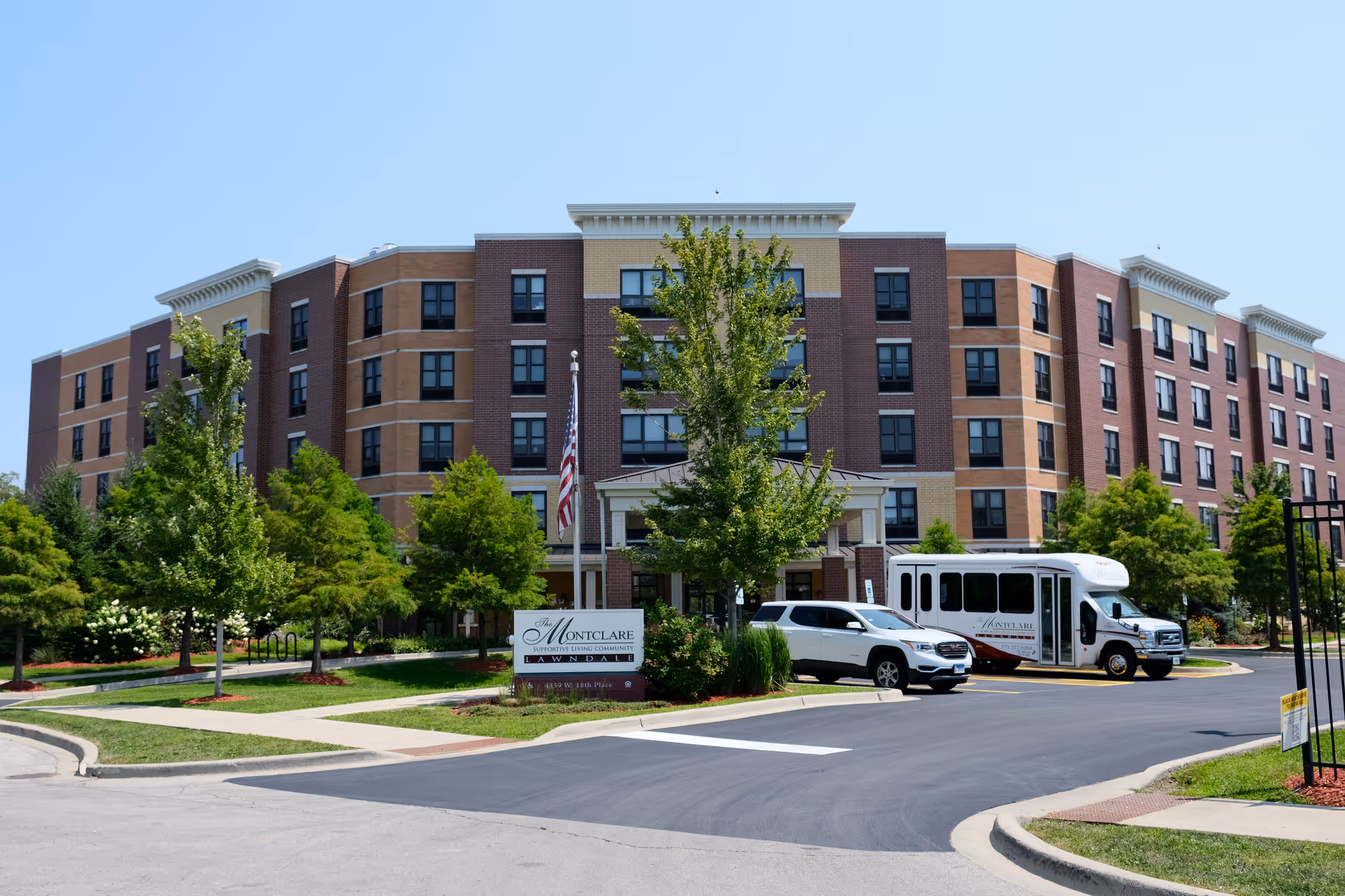 Exterior view of Montclare Supportive Living Community building with a multi-story brick facade, surrounded by green trees and landscaping. There is a driveway with a white SUV and a shuttle bus parked near the entrance. A sign in front reads 'The Montclare Supportive Living Community Lawndale'.