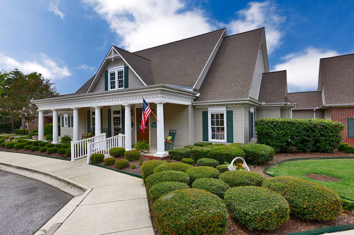 Exterior view of a single-story building with a steep roof and white columns supporting a covered porch. The building has beige siding with green shutters on the windows. There is a neatly trimmed garden with bushes and a small American flag hanging near the entrance. The sky is partly cloudy.