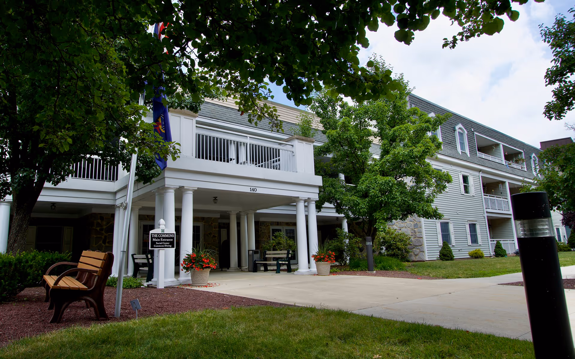 Exterior view of a senior living facility named Westminster Woods showing the main entrance area called The Commons. The building has white columns supporting a covered porch with benches and potted plants with red flowers. There are trees and green grass surrounding the paved walkway leading to the entrance.