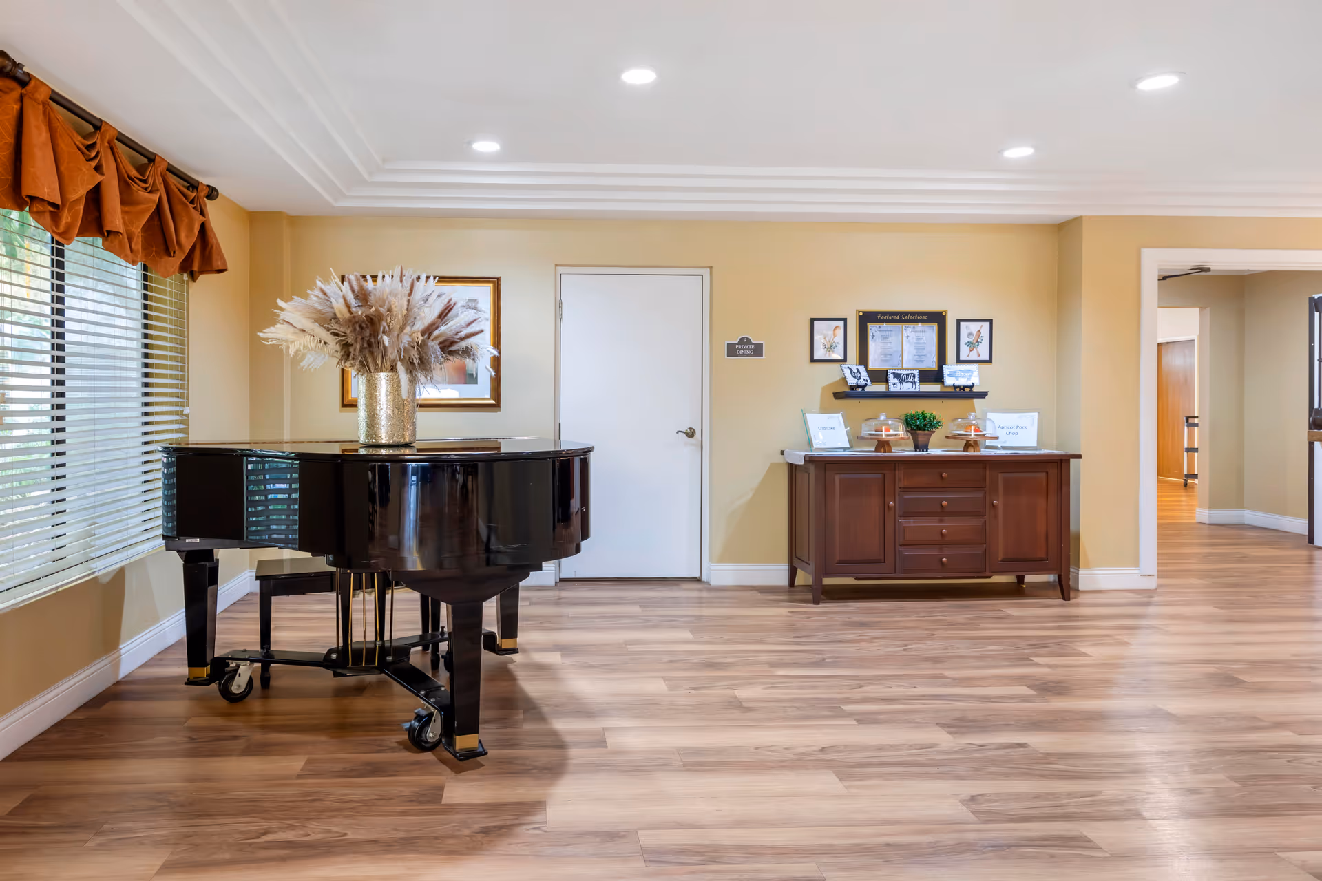 A bright room with light wood flooring and cream-colored walls featuring a black grand piano with a vase of dried flowers on top near a window with brown valance curtains. On the opposite wall, there is a wooden sideboard with framed pictures, informational signs, and a small potted plant. A white door is centered on the wall behind the sideboard, and an open doorway leads to another room on the right.
