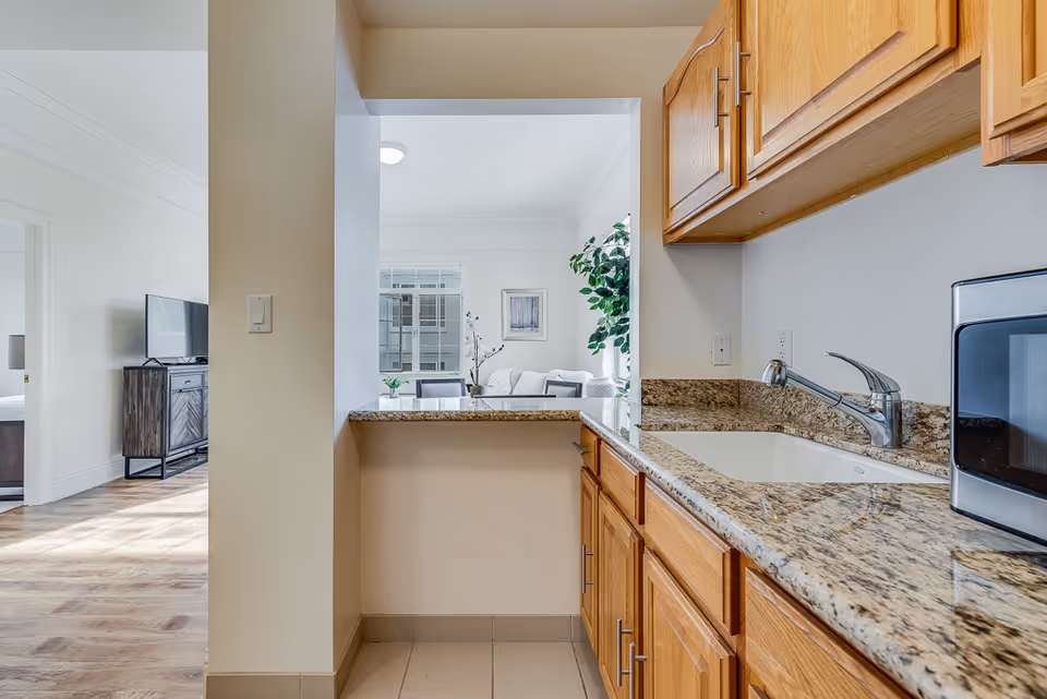 View of a kitchen area with wooden cabinets, granite countertops, a sink, and a microwave on the right side. Through a pass-through window, a living room with a sofa, a plant, and a framed picture on the wall is visible. To the left, there is a glimpse of a bedroom with a TV on a stand.