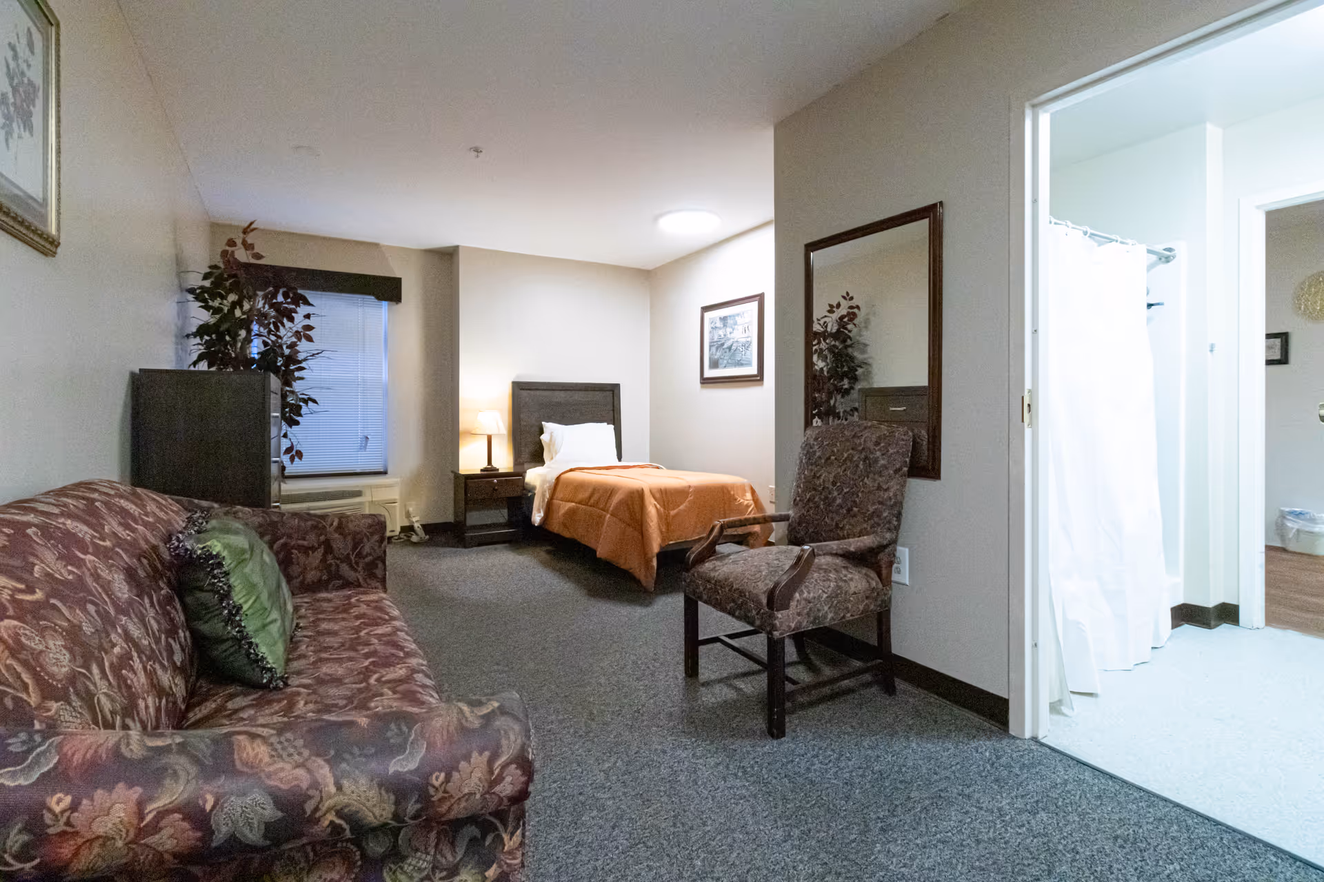 Interior view of a senior living facility room at Somerset Court of University Place featuring a single bed with an orange blanket, a nightstand with a lamp, a patterned armchair, a floral-patterned sofa with a green pillow, a large mirror on the wall, a potted plant, and a bathroom with a white shower curtain visible through an open door.
