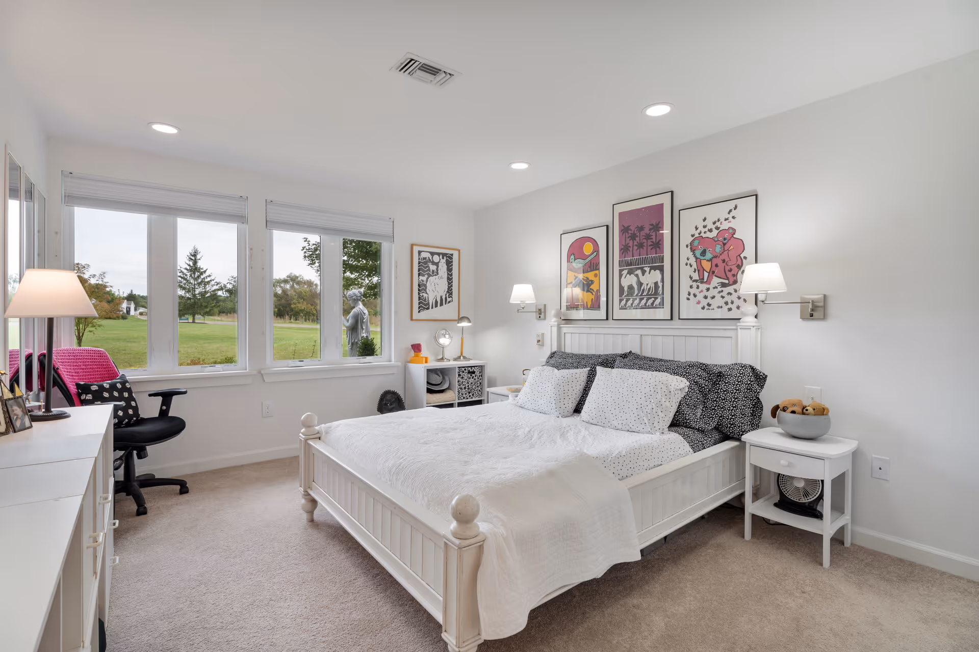 A bright and cozy bedroom with a white wooden bed frame and white bedding accented by black and white pillows. Three colorful framed artworks hang above the bed. There are two wall-mounted lamps on either side of the bed. A white nightstand with a bowl of stuffed animals and a small fan is next to the bed. Across from the bed is a white dresser with a lamp and framed photos. A black office chair with a pink blanket is positioned near three large windows that offer a view of a green lawn and trees outside. The room has beige carpet and white walls with recessed ceiling lights.