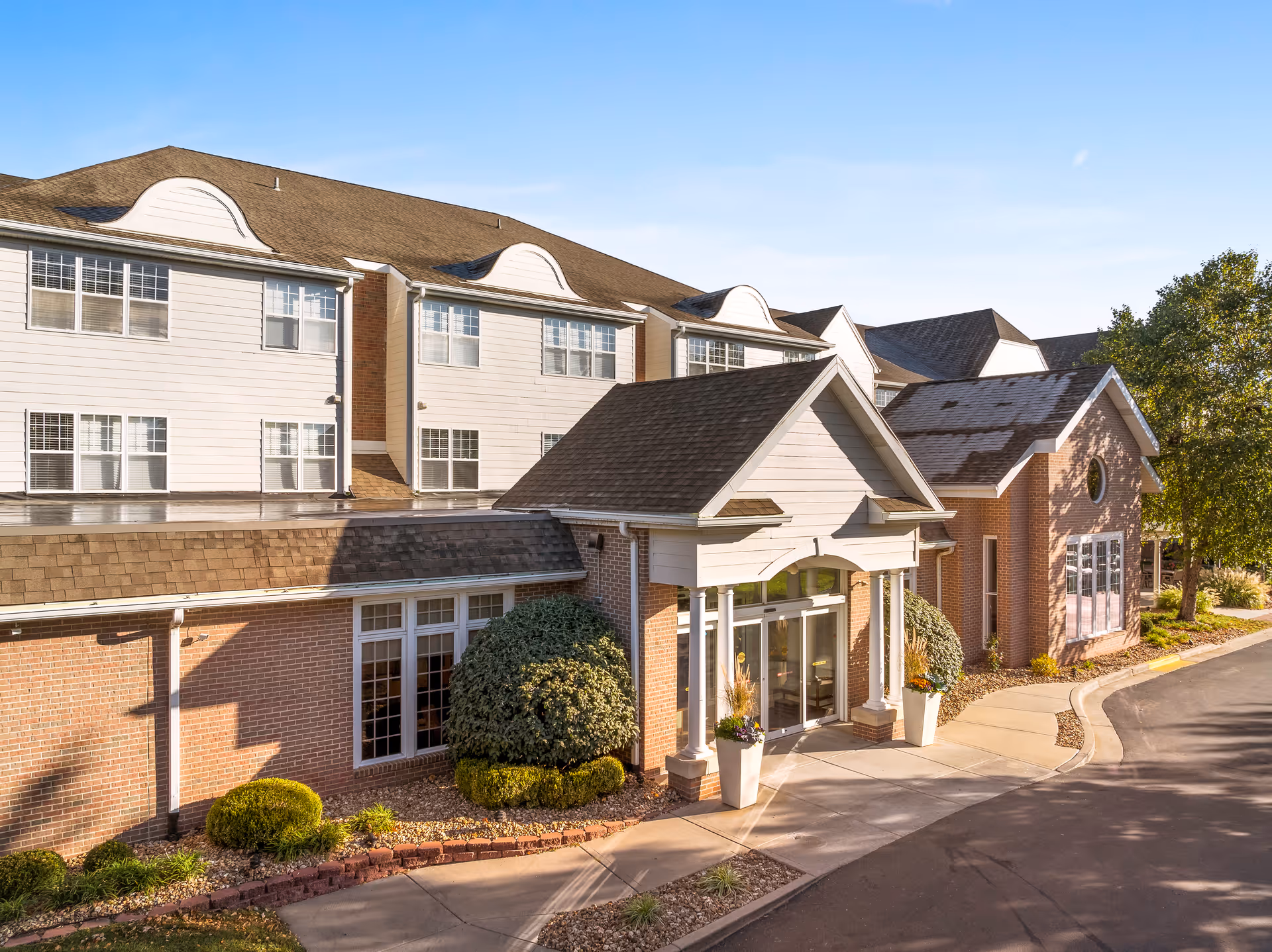 Front entrance and facade of a multi-story brick senior living building with a covered portico, landscaped beds, and a driveway.