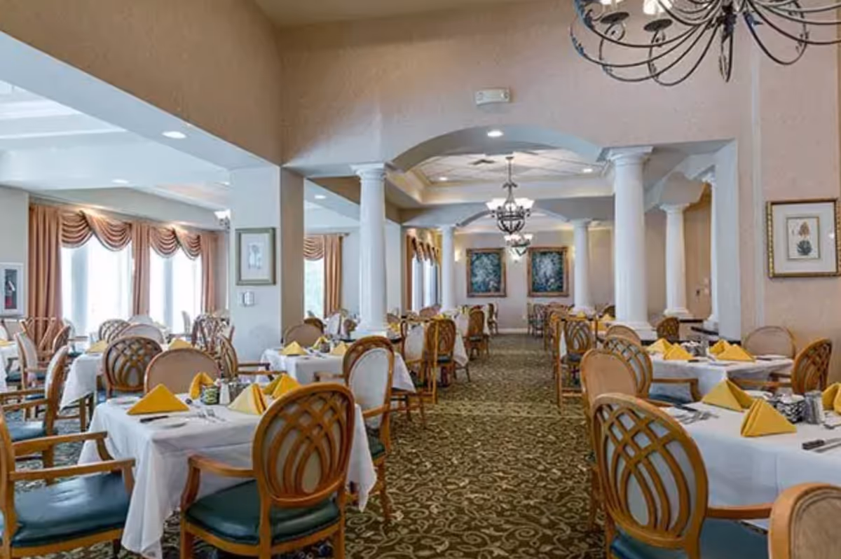Large formal dining room with tables set with white tablecloths and yellow napkins, wooden chairs, chandeliers, and decorative columns.
