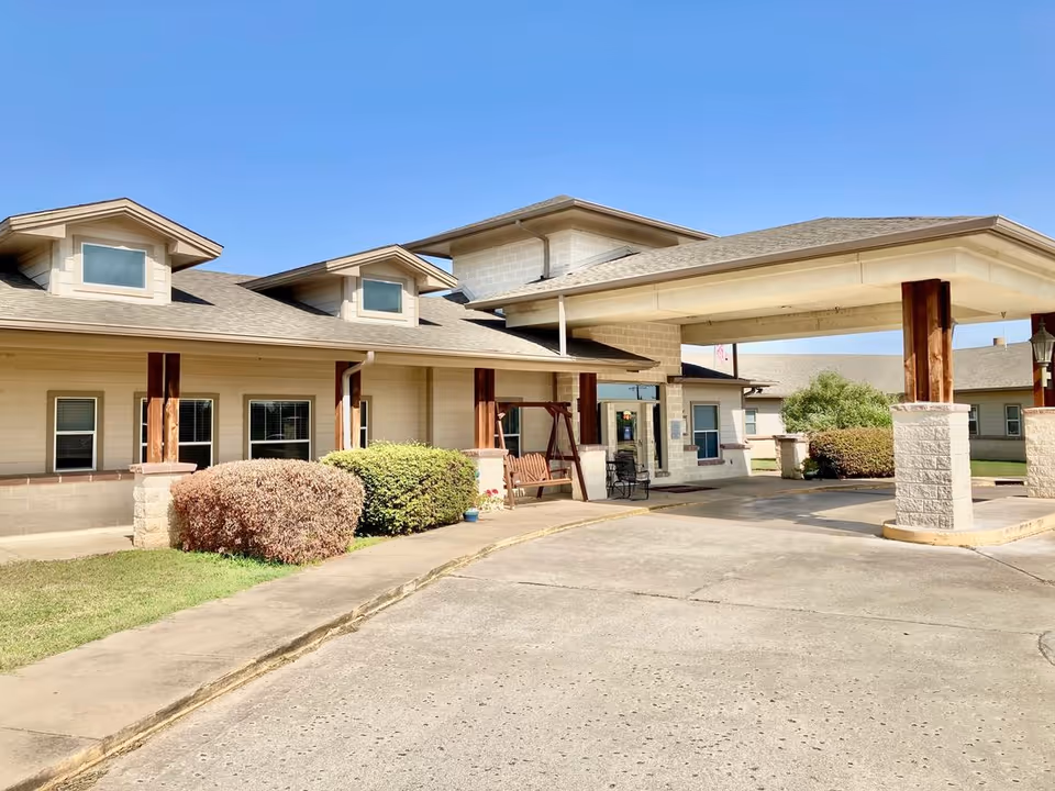 Front entrance of Windchime at the Village Assisted Living with a covered porte-cochère, bench swing, shrubs, and building facade under a clear blue sky.