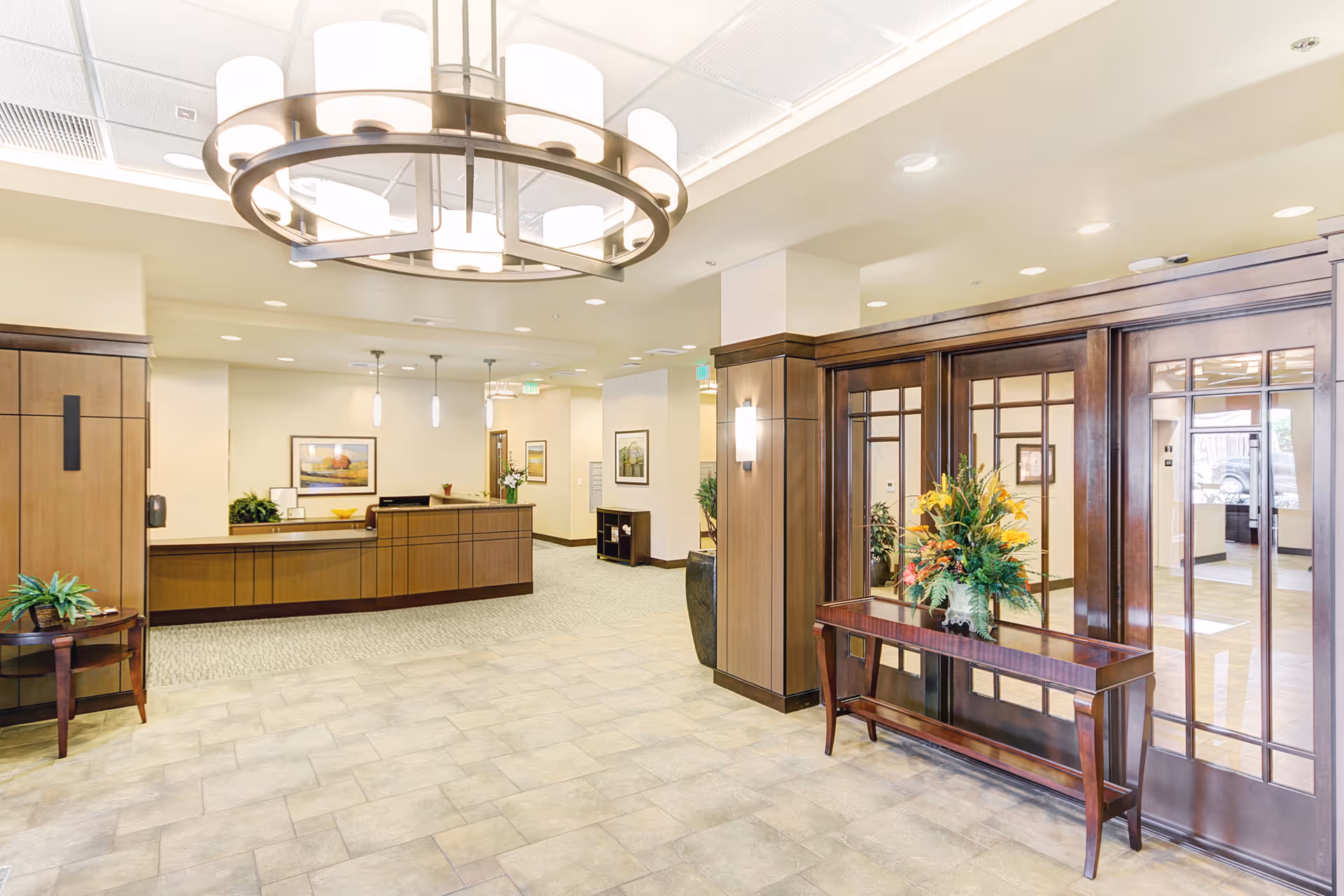 Bright and spacious reception area with a wooden front desk, decorative plants, framed artwork on the walls, a large circular chandelier hanging from the ceiling, and a wooden table with a floral arrangement in front of glass-paneled wooden doors.
