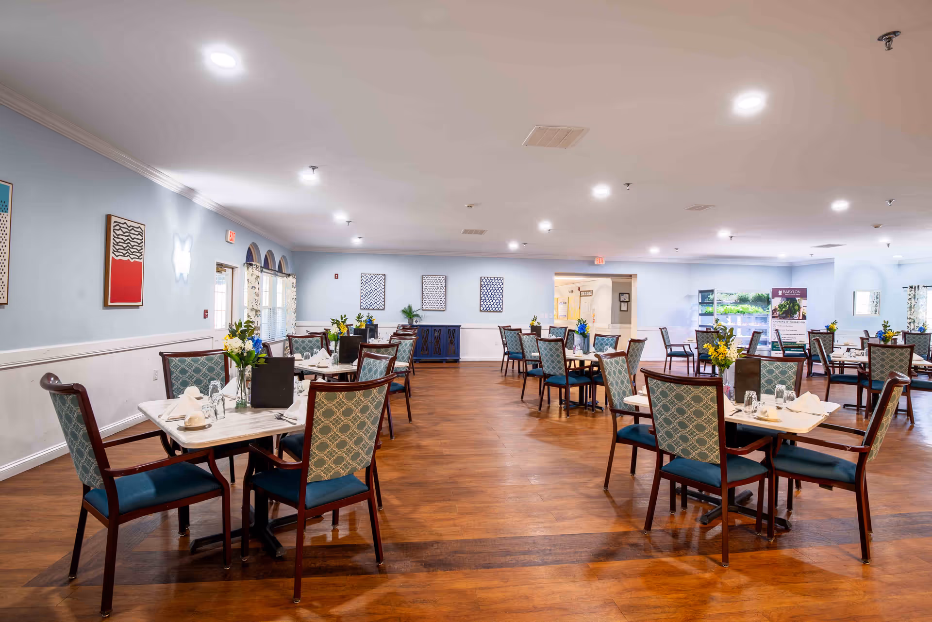 A spacious dining room with multiple tables and chairs arranged neatly. Each table is set with white napkins, glasses, and small flower vases. The room has light blue walls adorned with framed artwork and large windows with patterned curtains. The floor is wooden, and the ceiling has recessed lighting.