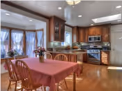 A bright kitchen and dining area with wooden floors and cabinetry. The dining table is covered with a red tablecloth and surrounded by wooden chairs. The kitchen features stainless steel appliances including a microwave and stove, with a window above the sink letting in natural light. There are decorative plants and flowers on the table and counters.