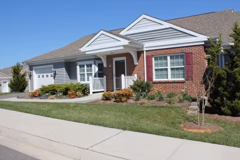 Exterior view of a single-story residential building with a brick and siding facade, a small front porch with white railings, a garage, and well-maintained landscaping including bushes and a small tree.