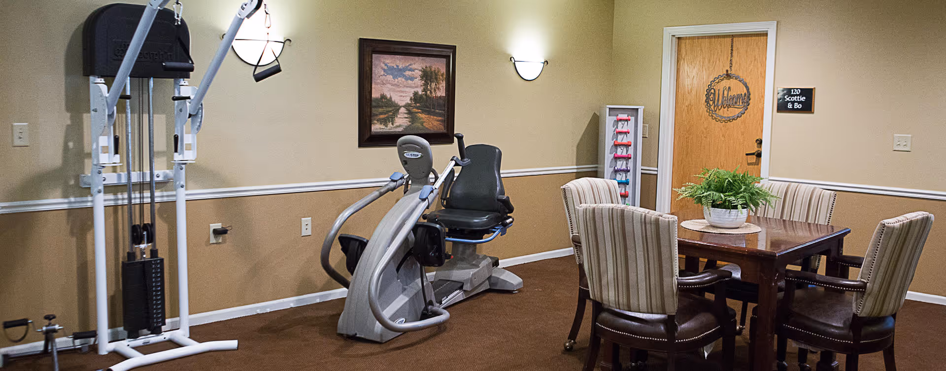 A room with exercise equipment including a weight machine and a recumbent bike on the left side, and a wooden table with four striped upholstered chairs on the right side. There is a potted plant on the table, a painting on the wall, and a door with a 'Welcome' sign and a plaque that reads '120 Scottie & Bo'.