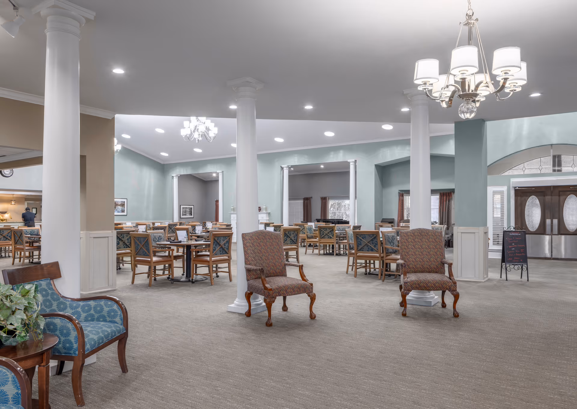 Spacious dining room with multiple wooden tables and chairs arranged neatly. The room features large white columns, soft green walls, carpeted floor, and elegant chandeliers hanging from the ceiling. There are two upholstered armchairs in the foreground and a small table with a plant on the left side. Double doors with oval glass panels are visible in the background.