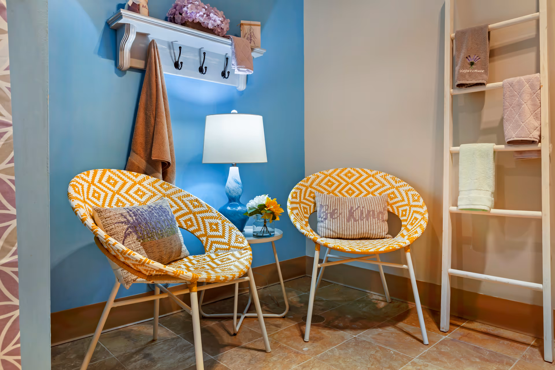 A cozy corner with two yellow and white patterned chairs, each with a decorative pillow. Between the chairs is a small round table with a blue lamp and a small flower arrangement. On the wall above one chair is a white shelf with hooks holding towels, and to the right is a wooden ladder rack with more towels hanging on it. The walls are painted blue and beige, and the floor is tiled.