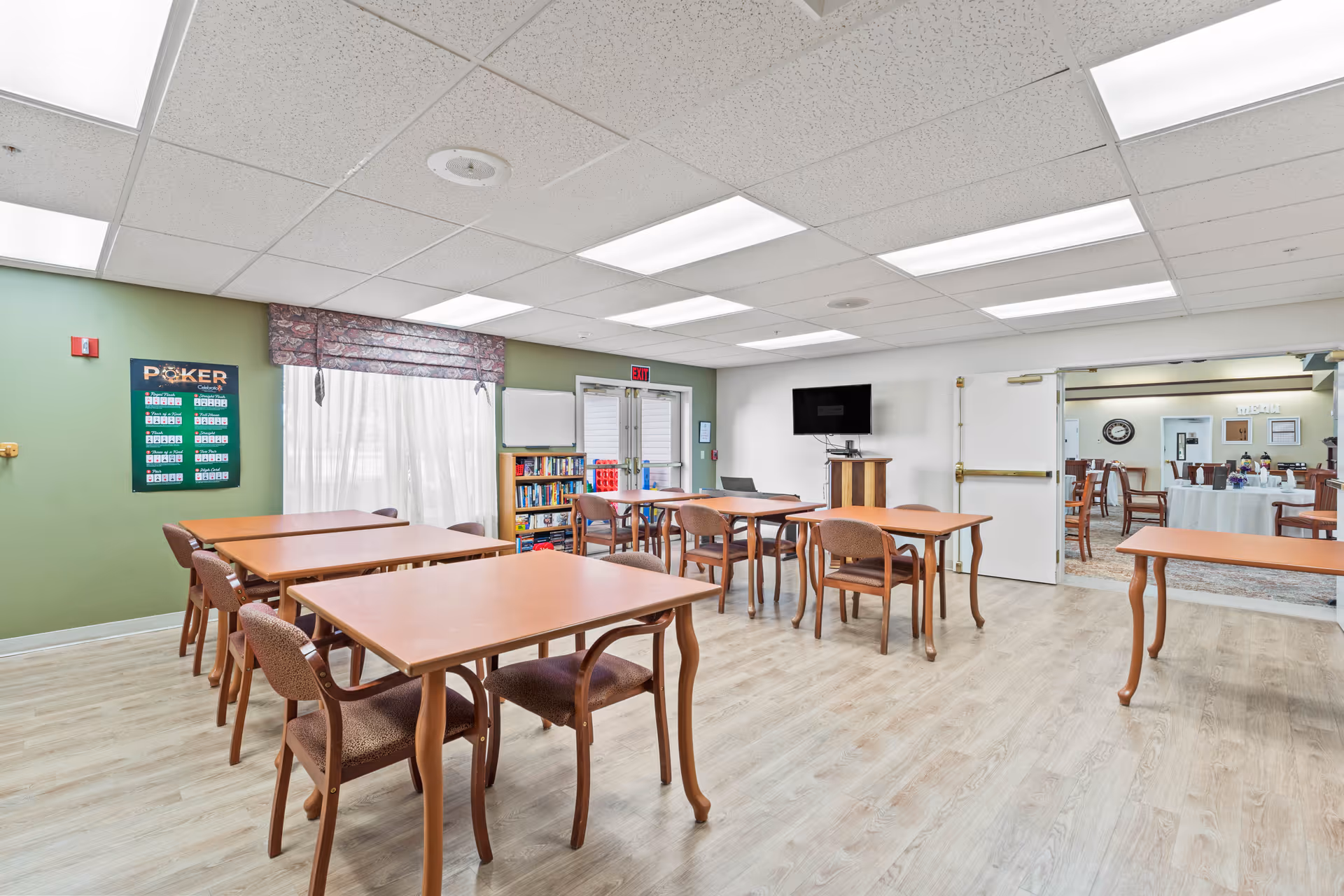 A bright room with several wooden tables and chairs arranged neatly on a light wood floor. The walls are painted green and white, with a window covered by sheer curtains and a valance. A bookshelf filled with books and games is visible near the window. A TV is mounted on the wall above a wooden stand. Double doors lead to another room with round tables and chairs set up, suggesting a dining or activity area.
